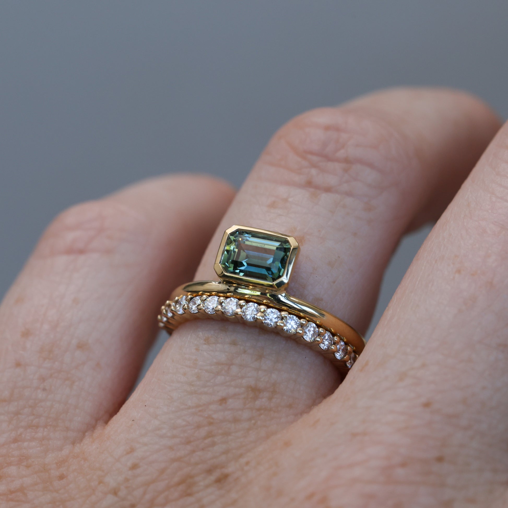 0.84ct emerald cut australian parti sapphire offset bezel engagement ring on hand with wedding band